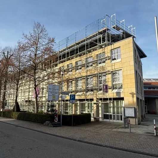 Ein mehrstöckiges Gebäude mit gelber Fassade wird von einem Gerüst umgeben, Bäume sind kahl.